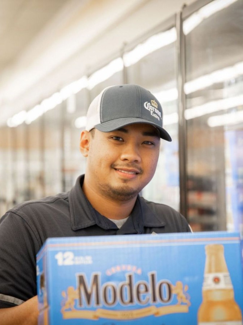 Young man smiles at the camera while stocking cold case with Modelo beer