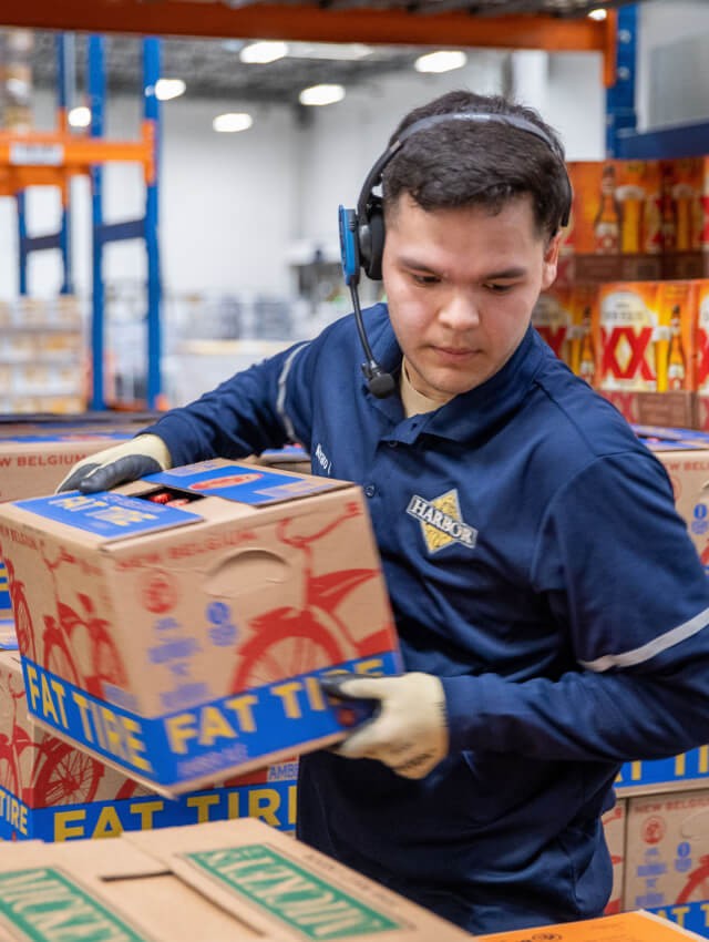 Young man moves a case of beer in warehouse wearing work gloves and a Harbor Distributing shirt